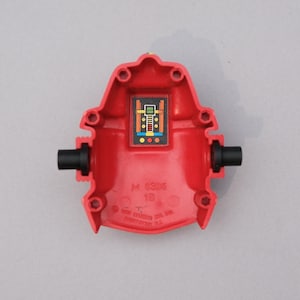 May include: A red plastic toy component with a rectangular, colorful graphic in the center. Two black cylindrical pieces protrude from the sides. The component has the text "M 3395 1B" and "© HASBRO INC. 1985 HASBRO IND. LTD."