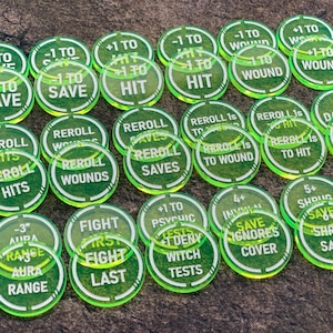 Puede incluir: Una colección de fichas de juego verdes transparentes con texto blanco. Las fichas tienen varias frases como "+1 to Save", "Reroll Hits" y "Attack". Estas fichas están diseñadas para juegos de mesa.