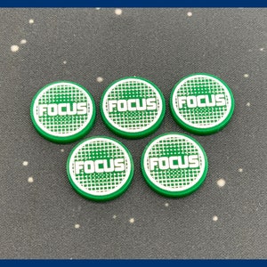 May include: Five green and white circular tokens with the word "FOCUS" in white block letters. The tokens have a green grid pattern in the center and a green border. The tokens are arranged on a dark gray surface.
