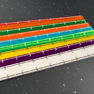 May include: A stack of colorful rulers in red, orange, teal, green, yellow, purple, and white. Each ruler is marked with inch measurements from 1 to 9, with a white border and black markings. The rulers are stacked on a dark speckled surface.