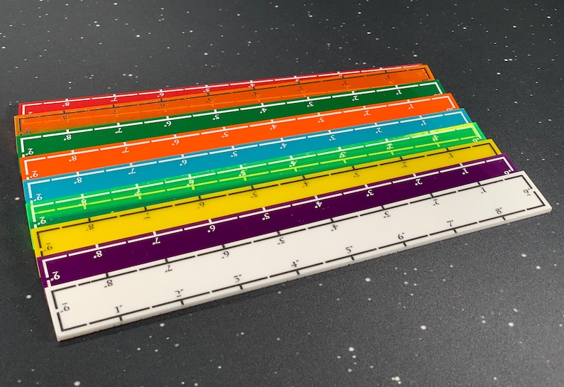 May include: A stack of colorful rulers in red, orange, teal, green, yellow, purple, and white. Each ruler is marked with inch measurements from 1 to 9, with a white border and black markings. The rulers are stacked on a dark speckled surface.
