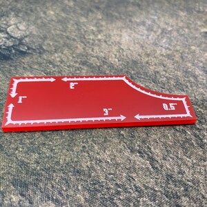 May include: Red plastic sewing template with white markings. The template is shaped like a curved rectangle with a rounded corner. The markings include measurements in inches: 1", 2", 3", and 0.5".