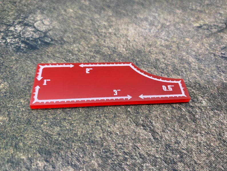 May include: Red plastic sewing template with white markings. The template is shaped like a curved rectangle with a rounded corner. The markings include measurements in inches: 1", 2", 3", and 0.5".