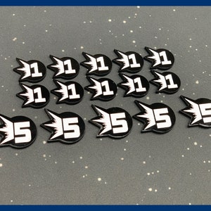 May include: A collection of black and white game tokens. The tokens are shaped like a burst with a number in the center. Some tokens have the number 1, while others have the number 5. The tokens are arranged on a gray surface.