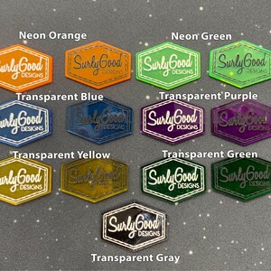 May include: A collection of hexagonal acrylic stickers with the text "Surly Good Designs" in white lettering. The stickers are in various colours, including neon orange, neon green, transparent blue, transparent purple, transparent yellow, transparent green, and transparent grey.