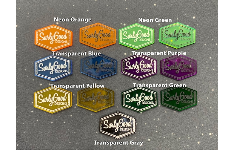 May include: A collection of hexagonal acrylic stickers with the text "Surly Good Designs" in white lettering. The stickers are in various colours, including neon orange, neon green, transparent blue, transparent purple, transparent yellow, transparent green, and transparent grey.