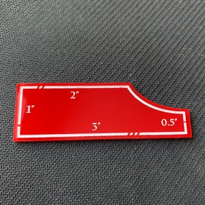 Pode incluir: Um modelo de pl&aacute;stico vermelho com marcas brancas para cortar tecido. O modelo tem 7,6 cm de comprimento, 5,1 cm de largura e uma curva de 1,3 cm.