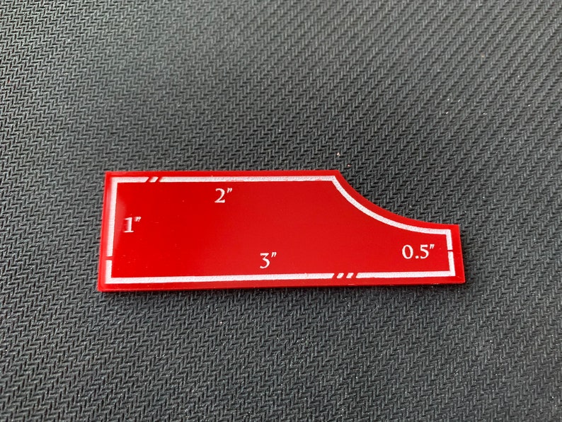 Pode incluir: Um modelo de pl&aacute;stico vermelho com marcas brancas para cortar tecido. O modelo tem 7,6 cm de comprimento, 5,1 cm de largura e uma curva de 1,3 cm.