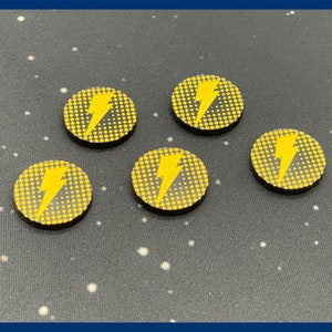 May include: Five yellow and black circular game tokens with a lightning bolt design. Each token has a yellow lightning bolt on a yellow dotted background. The tokens are on a dark gray surface with white dots.