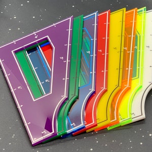 K&ouml;nnte beinhalten: Ein Satz von sieben bunten Plastiklinealen mit verschiedenen Markierungen und Formen. Die Lineale sind f&auml;cherf&ouml;rmig angeordnet, wobei das violette Lineal oben und das gelbe Lineal unten liegt. Die Lineale sind alle transparent und haben wei&szlig;e Markierungen.