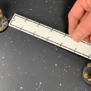 May include: A white ruler with black markings is held near two miniature figurines on a dark surface. The figurines are of fantasy characters, one a large, muscular figure and the other a warrior in red and gold armor. The ruler is marked in inches.