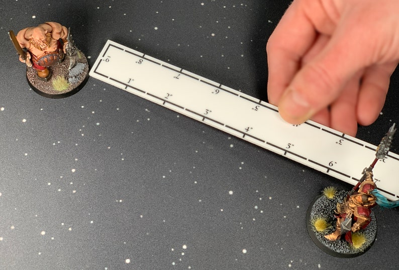 May include: A white ruler with black markings is held near two miniature figurines on a dark surface. The figurines are of fantasy characters, one a large, muscular figure and the other a warrior in red and gold armor. The ruler is marked in inches.