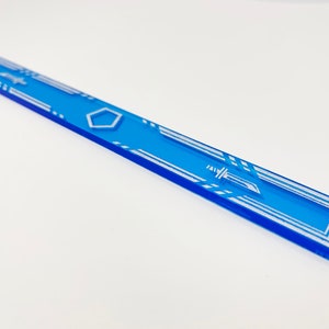K&ouml;nnte beinhalten: Ein blauer Acryl-Lineal mit einem wei&szlig;en geometrischen Muster und einer Schwertdarstellung. Das Lineal ist etwa 30 cm lang.