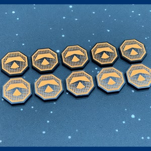 May include: Ten black and orange game tokens arranged in two rows. Each token is an octagon shape with an orange design of a stylized arrow and grid pattern. The tokens are on a dark blue surface with white specks.