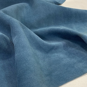 Könnte beinhalten: Nahaufnahme eines Stücks blauen Wildleder-Stoffs. Das Material ist weich und hat eine strukturierte Oberfläche. Der Stoff ist gefaltet und drapiert, wodurch der natürliche Fluss und die Textur des Materials sichtbar werden. Die Farbe ist ein gedämpftes, staubiges Blau.