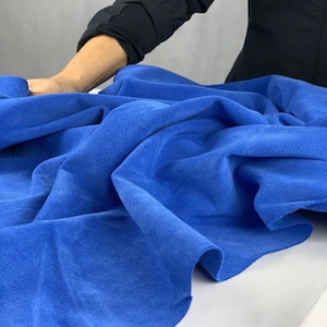 Könnte beinhalten: Ein leuchtend blauer Stoff mit einer weichen, Wildleder-ähnlichen Textur ist über eine Oberfläche drapiert. Der Stoff ist in sanften Falten angeordnet. Arm und Hand einer Person sind sichtbar und berühren das Material sanft. Der Hintergrund ist neutralgrau.
