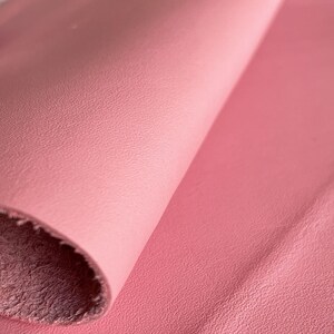 May include: Close-up of a soft, pink leather fabric with a textured surface. The fabric is folded over, revealing the underside.