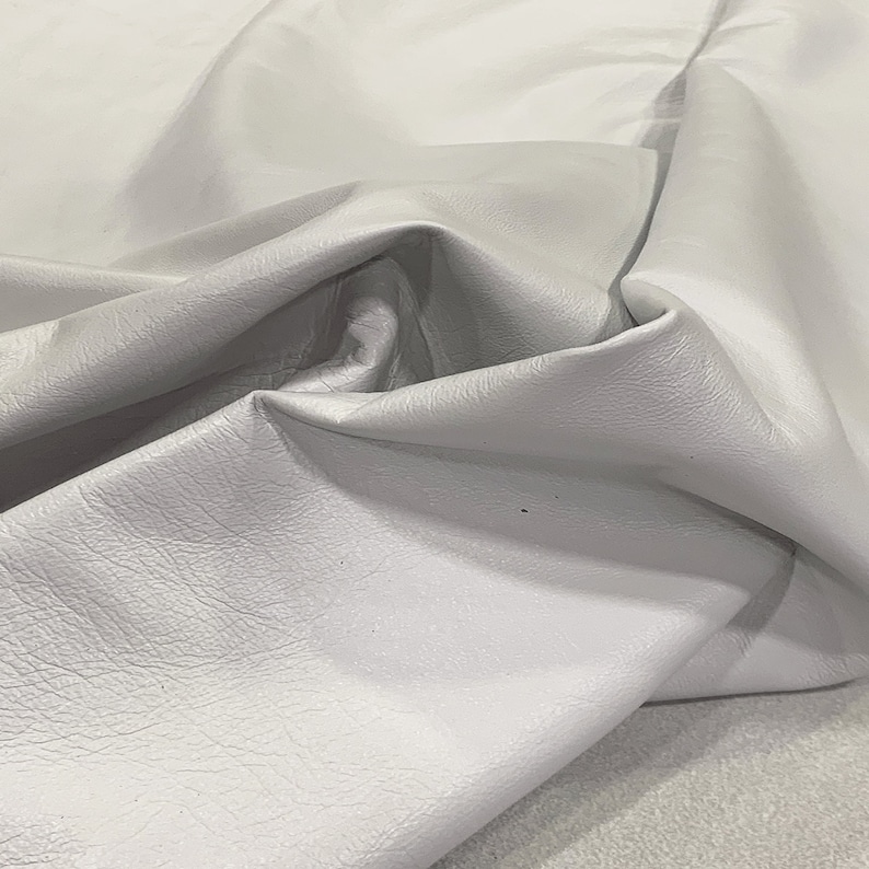May include: A close-up of a piece of white leather with a textured surface. The leather is folded and draped, showing its soft and supple texture.