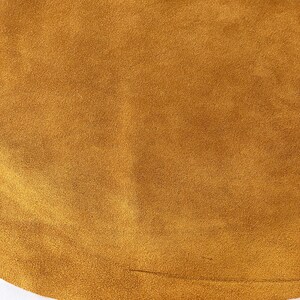 May include: Close-up of a textured, golden-brown suede fabric. The material has a soft, velvety appearance with a subtle sheen. The color is a warm, earthy tone, and the fabric appears to be smooth to the touch. The image is well-lit, highlighting the texture and color.