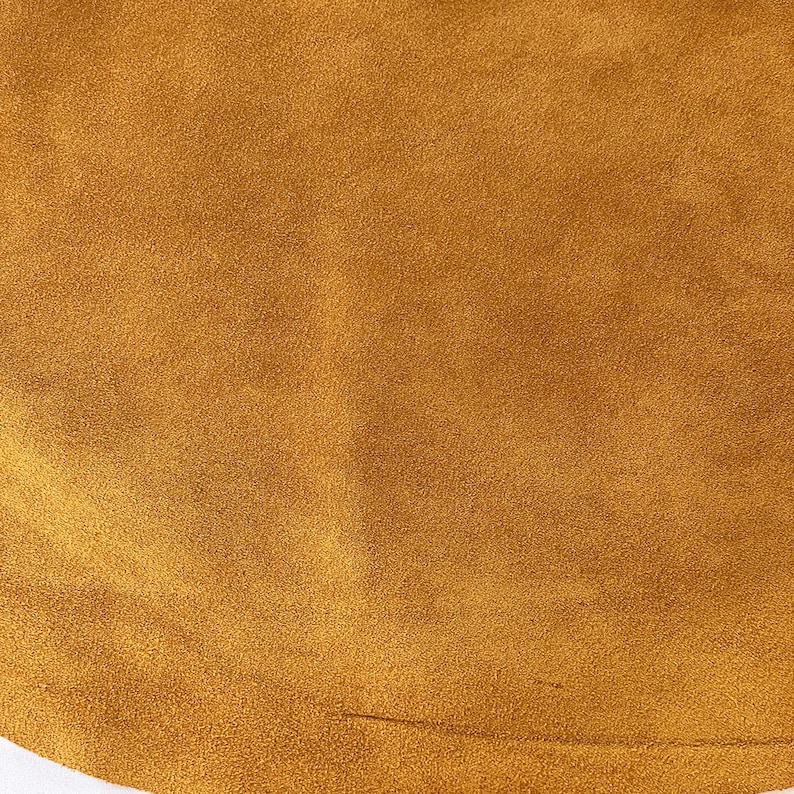 May include: Close-up of a textured, golden-brown suede fabric. The material has a soft, velvety appearance with a subtle sheen. The color is a warm, earthy tone, and the fabric appears to be smooth to the touch. The image is well-lit, highlighting the texture and color.