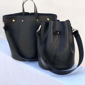 May include: Two black leather tote bags. The larger bag has a top handle and a wide, flat base. The smaller bag has a drawstring closure and a long shoulder strap.