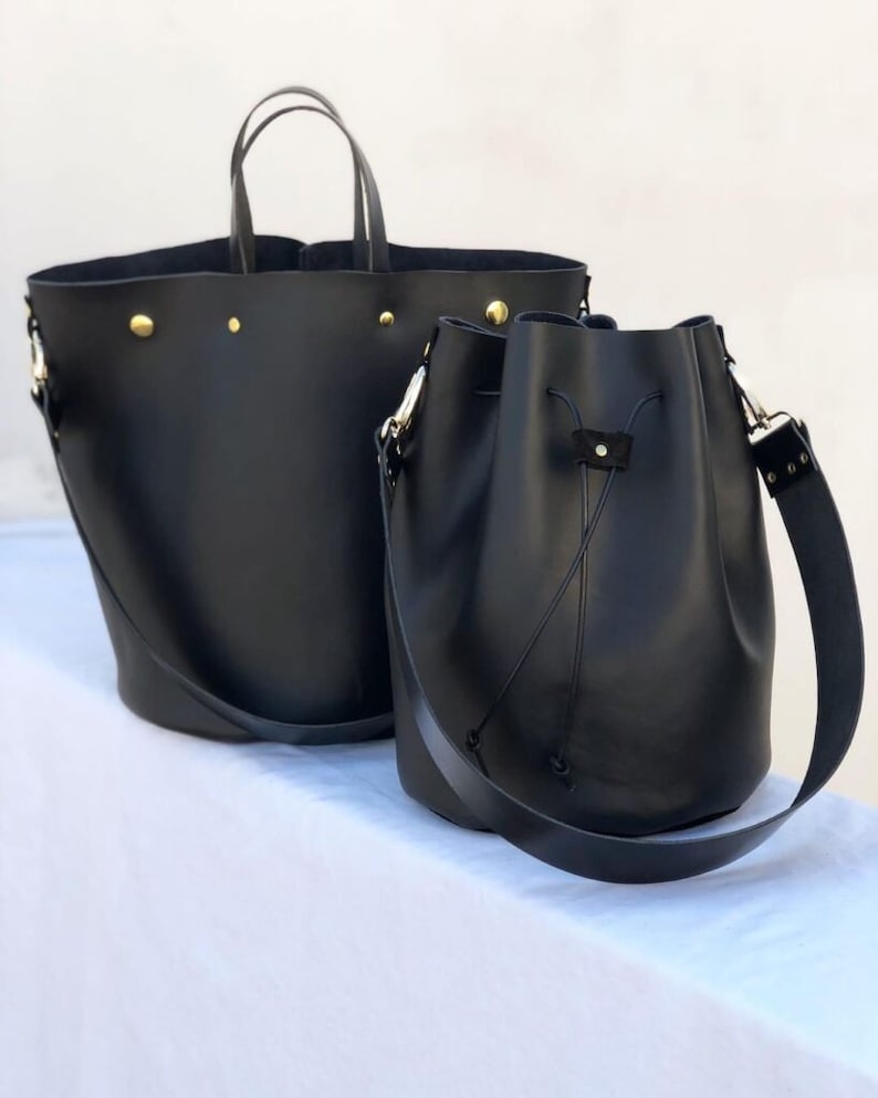 May include: Two black leather tote bags. The larger bag has a top handle and a wide, flat base. The smaller bag has a drawstring closure and a long shoulder strap.