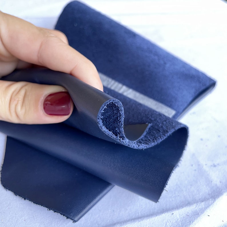 Pode incluir: Close-up de folhas de couro azul marinho, com uma se&ccedil;&atilde;o transversal vis&iacute;vel revelando a textura. O couro &eacute; liso e parece ser de alta qualidade. Um peda&ccedil;o de camur&ccedil;a azul marinho est&aacute; no fundo.