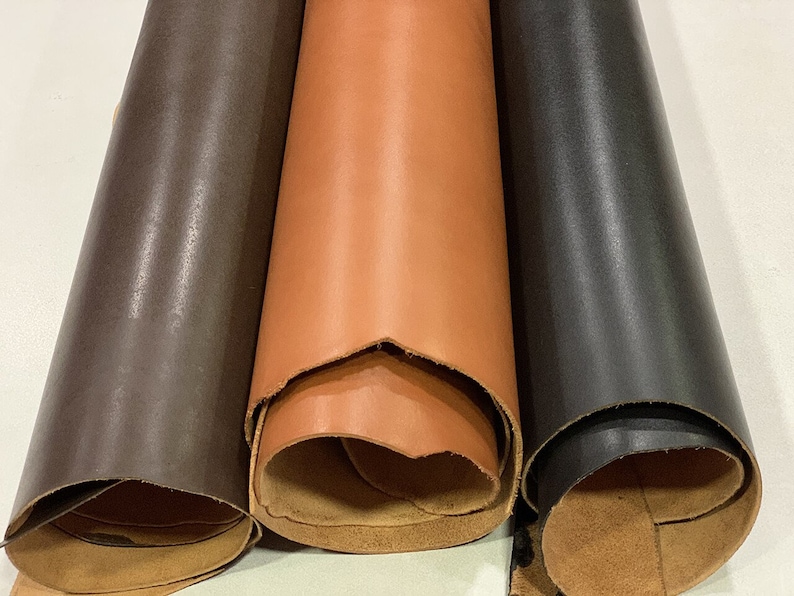 K&ouml;nnte beinhalten: Drei Rollen Leder in verschiedenen Farben: Braun, Orange und Schwarz. Das Leder ist aufgerollt und die Kanten sind sichtbar.