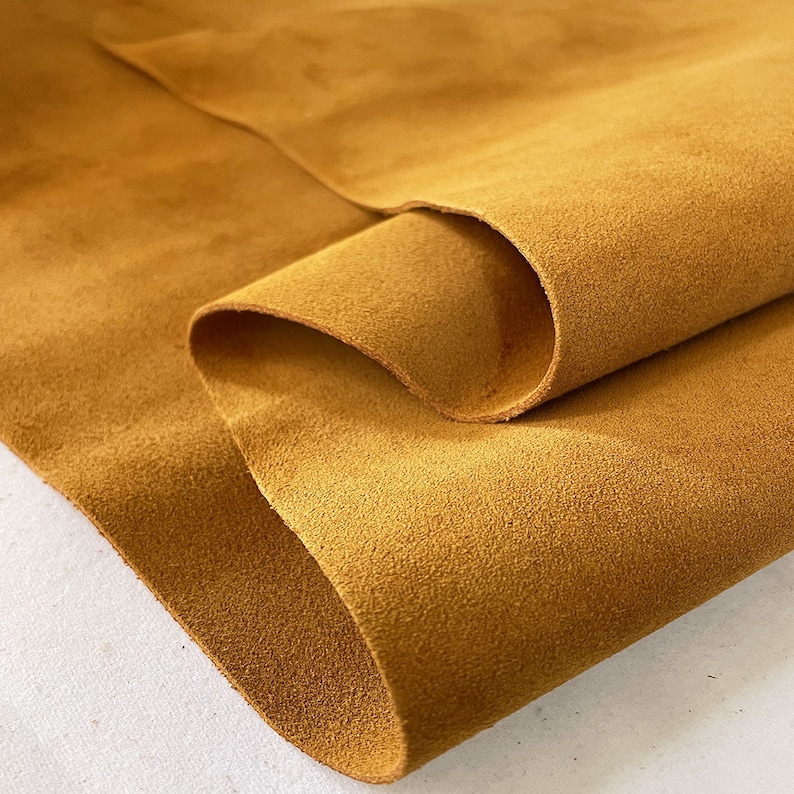 May include: Close-up of a piece of mustard-colored suede leather. The leather is folded over, revealing its soft, textured surface. The color is a warm, earthy tone, ideal for crafting or fashion projects.
