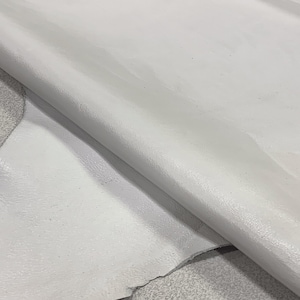 May include: A close-up of a piece of white leather. The leather is smooth and has a slight sheen. It is folded in half, showing the inside of the leather.