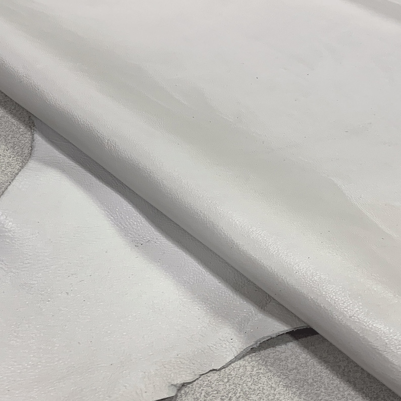 May include: A close-up of a piece of white leather. The leather is smooth and has a slight sheen. It is folded in half, showing the inside of the leather.