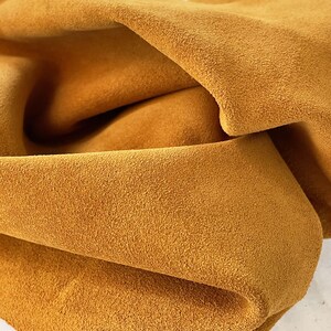 May include: Close-up of a folded piece of mustard-colored suede leather. The leather has a soft, textured surface and a warm, inviting hue. The folds create shadows and depth, highlighting the material's flexibility. Ideal for crafting projects.