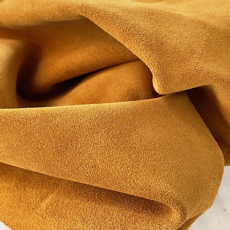 May include: Close-up of a folded piece of mustard-colored suede leather. The leather has a soft, textured surface and a warm, inviting hue. The folds create shadows and depth, highlighting the material's flexibility. Ideal for crafting projects.