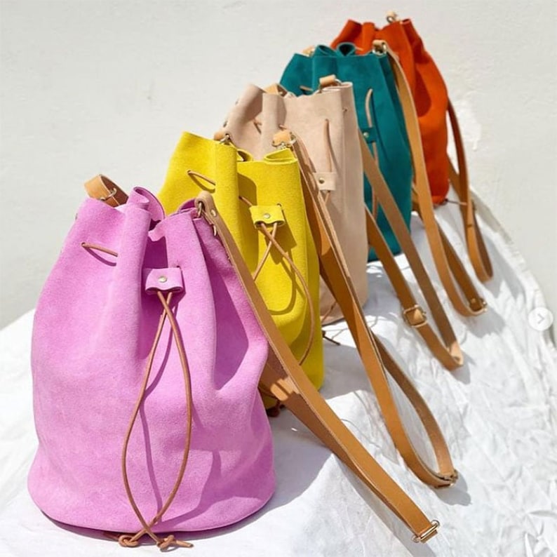 May include: A collection of colorful suede bucket bags in shades of pink, yellow, beige, teal, and orange. Each bag features a drawstring closure and a long, brown leather shoulder strap. The bags are displayed on a white surface.