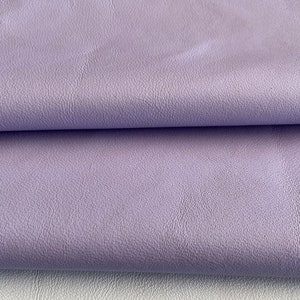 May include: Close-up of a soft, textured, lilac-colored leather fabric. The fabric is folded in half, showing the smooth, even texture.