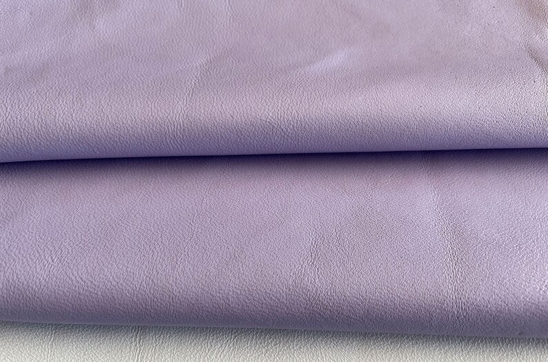 May include: Close-up of a soft, textured, lilac-colored leather fabric. The fabric is folded in half, showing the smooth, even texture.