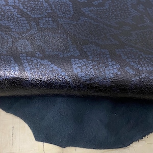 May include: A close-up of a dark blue leather fabric with a textured, embossed pattern. The fabric is folded over, revealing a smooth, dark blue suede underside.
