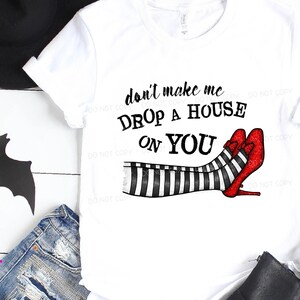 以下が含まれることがあります： 「don't make me DROP A HOUSE ON YOU」の文字が入った白いTシャツ。縞模様の脚と赤いグリッターヒールが特徴で、魔女をテーマにしています。高解像度のプリントファイルです。