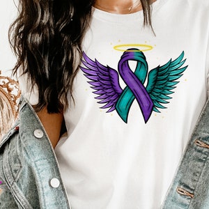 Puede incluir: Una camiseta blanca con una cinta morada y turquesa con alas de ángel y un halo. La cinta es un símbolo de concienciación para muchas causas.
