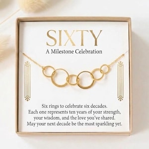 May include: A gold-toned necklace featuring six interconnected circular rings, presented in a square box. The card inside reads: SIXTY A Milestone Celebration. Six rings to celebrate six decades. Each one represents ten years of your strength, your wisdom, and the love you’ve shared. May your next decade be the most sparkling yet.