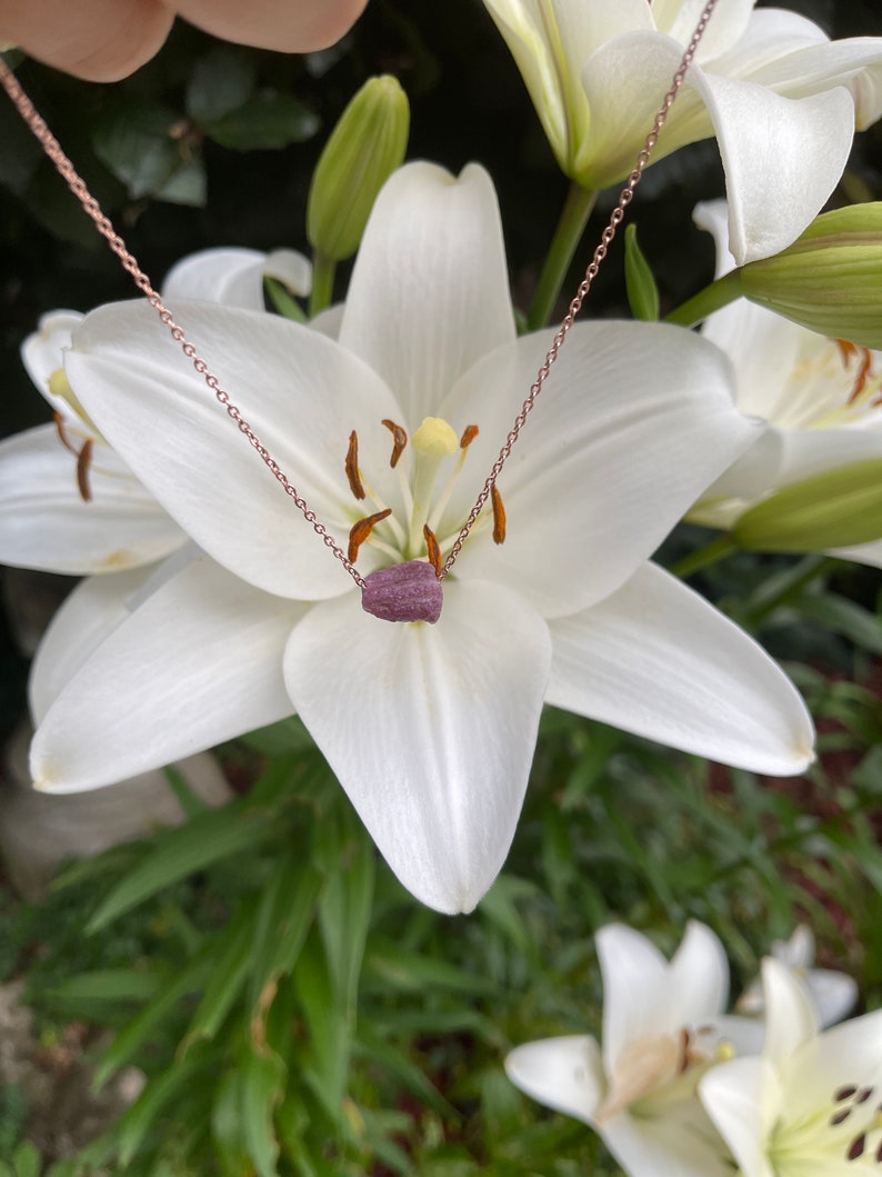 May include: A delicate rose gold chain necklace with a small, dark purple gemstone pendant. The necklace is displayed against a backdrop of white lilies.