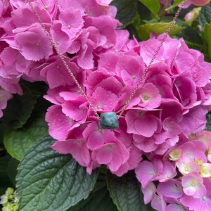 May include: A delicate gold chain necklace with a dark green gemstone pendant. The necklace is displayed against a backdrop of pink hydrangea flowers.