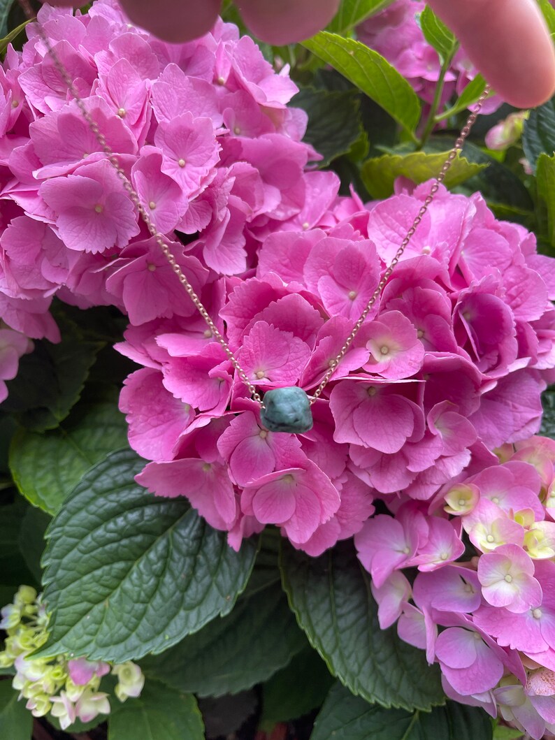 May include: A delicate gold chain necklace with a dark green gemstone pendant. The necklace is displayed against a backdrop of pink hydrangea flowers.