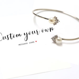 May include: Silver bangle bracelet with two pearl and crystal accents. The bracelet is on a white card with the text "Custom your own" and "Message Card".