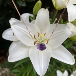 May include: A delicate rose gold chain necklace with a small, raw amethyst crystal pendant. The necklace is photographed against a backdrop of white lilies.