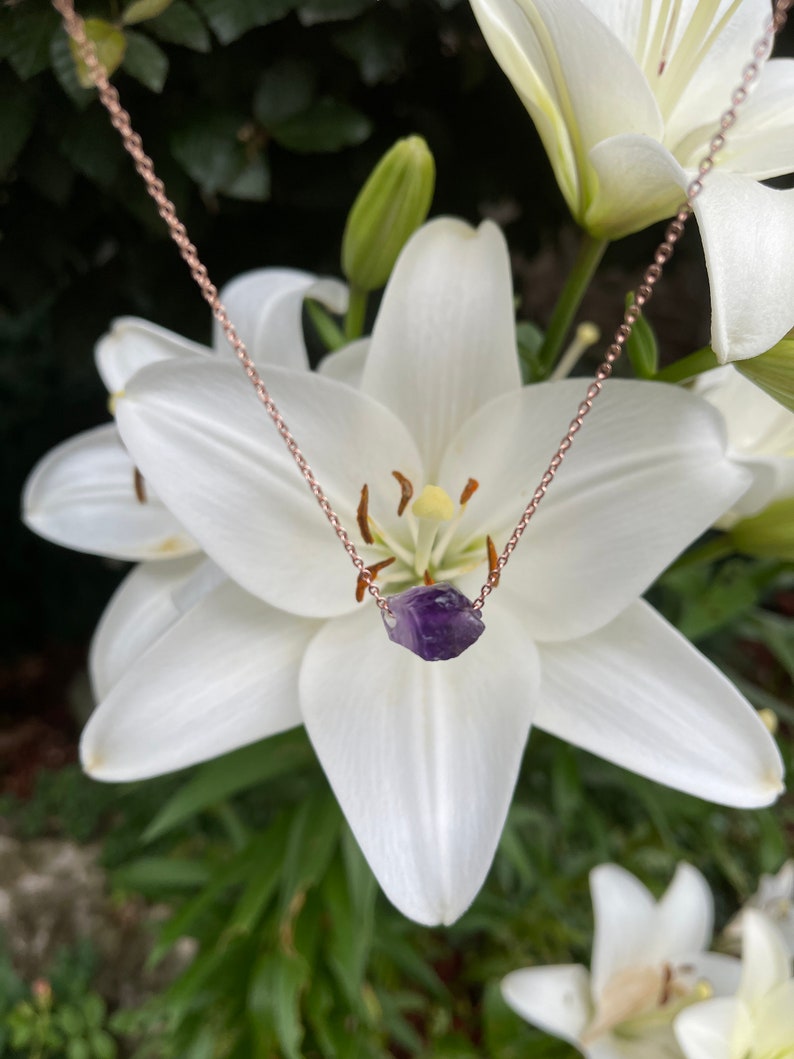 May include: A delicate rose gold chain necklace with a small, raw amethyst crystal pendant. The necklace is photographed against a backdrop of white lilies.