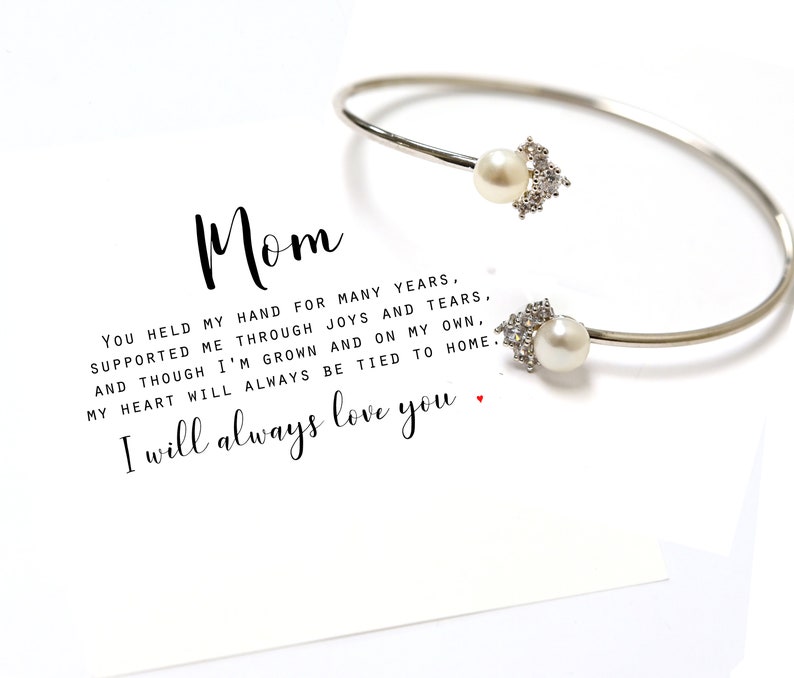 May include: A silver bangle bracelet with two pearl and crystal accents. The bracelet is accompanied by a card with the text "Mom You held my hand for many years. Supported me through joys and tears. And though I'm grown and on my own, my heart will always be tied to home. I will always love you."