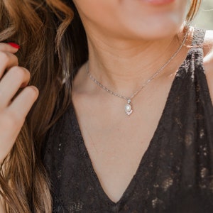 May include: A silver chain necklace with a pearl pendant. The pearl is surrounded by smaller clear stones.