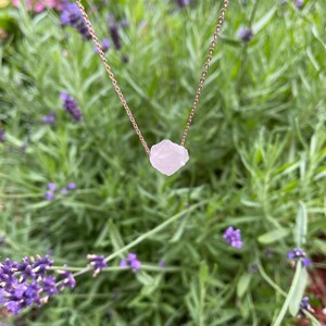 May include: A delicate rose gold chain necklace featuring a raw, natural, white quartz crystal pendant. The necklace is photographed against a backdrop of lavender flowers.