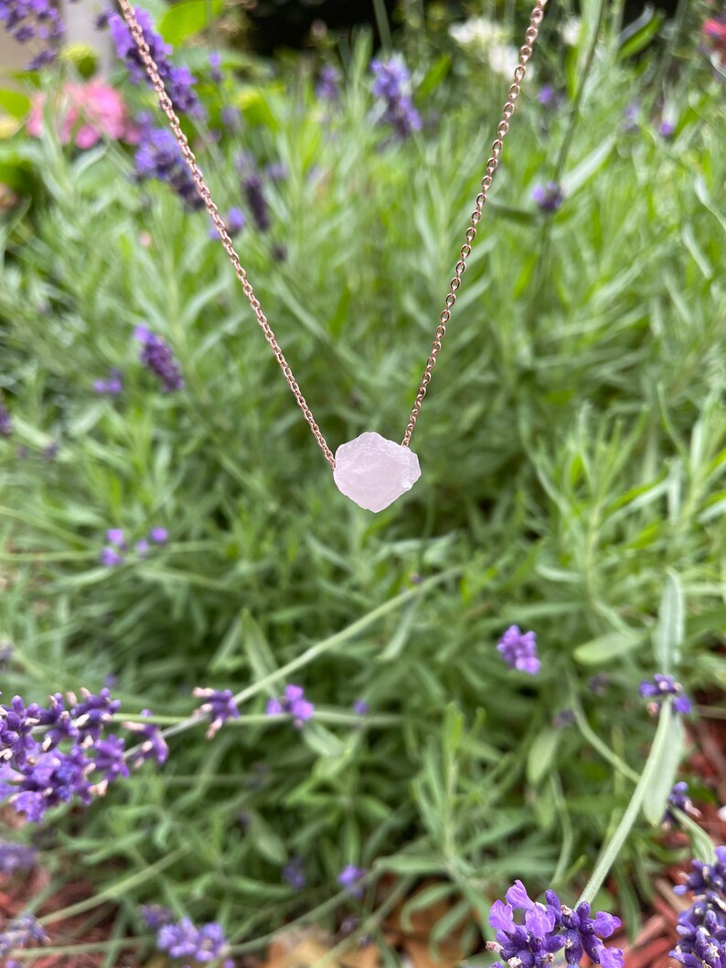 May include: A delicate rose gold chain necklace featuring a raw, natural, white quartz crystal pendant. The necklace is photographed against a backdrop of lavender flowers.